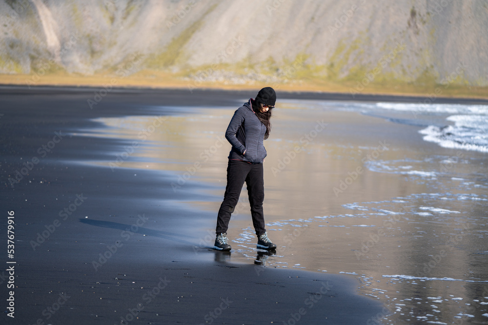portrait woman walking on the black volcanic sand beach with mountains in the background. She is wearing glasses and cap portrait woman walking on the black volcanic sand beach with mountains in the background. She is wearing glasses and cap