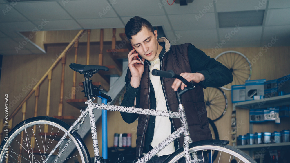 Attractive young man cheerful mechanic is busy talking on mobile phone and assembling bicycle handle-bar. People, maintenance and communication concept. Attractive young man cheerful mechanic is busy talking on mobile phone and assembling bicycle handle-bar. People, maintenance and communication concept.