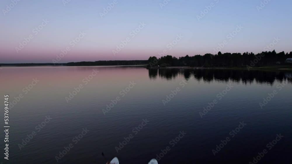 Aerial flyover  two young women paddling on a sup board on a lake towards the beach, healty activity, water sport. Calm lake surrounded by forests with a colorful summer sunset blue hour