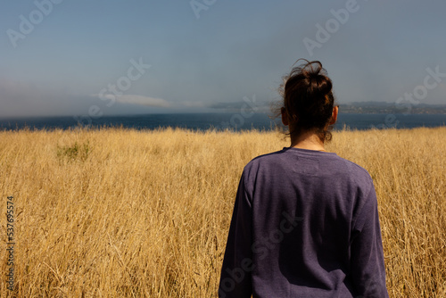 The girl from behind looks into the distance at the ocean, stands in a field with yellow grass, blue sky and fog. © Pablo Santos Somos