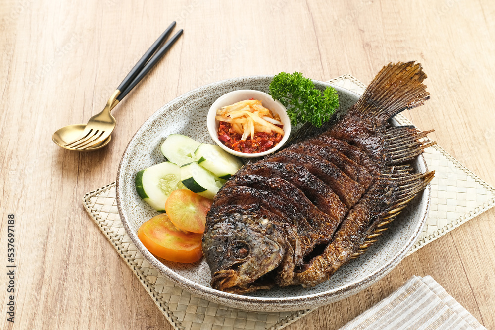 Gurame Bakar Madu, Grilled Gourami with honey and soy sauce. Served with sambal mangga (chilli sauce and grated mango), cucumber and tomatoes in white plate.
Gurame Bakar Madu, Grilled Gourami with honey and soy sauce. Served with sambal mangga (chilli sauce and grated mango), cucumber and tomatoes in white plate.