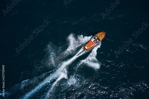 A luxurious wooden large boat with people is moving quickly on dark blue water making a white trail behind the boat. Wooden boat in motion on the water top view.