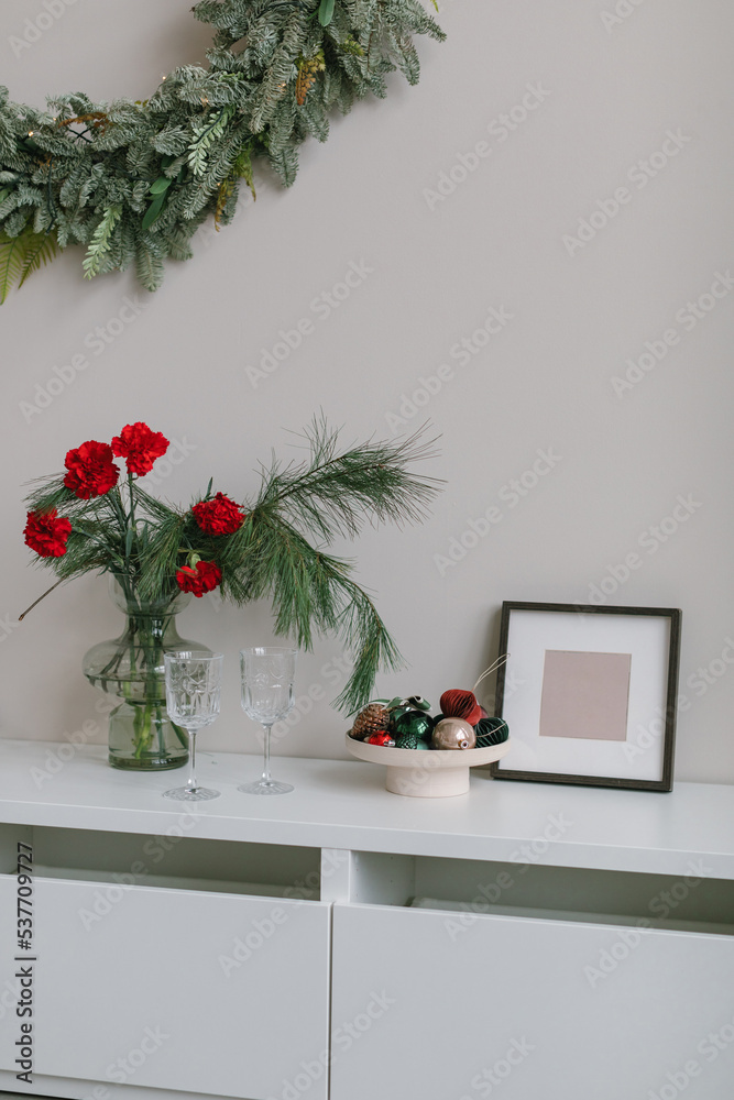 Christmas interior decor. Floral composition in stylish vase, Christmas toys in ceramic plate,  blank photo frame, glasses on  white dresser. Christmas wreath on the wall.