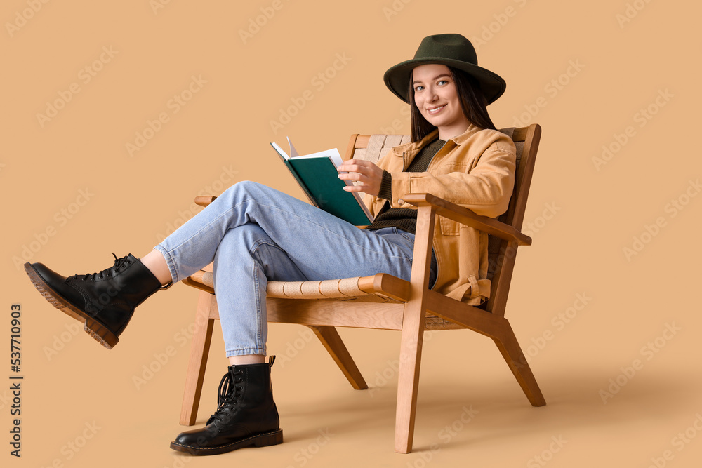 Young woman reading book in armchair on beige background