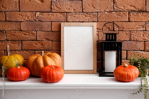 Blank frame with Halloween pumpkins, lantern and houseplant on mantelpiece near brick wall