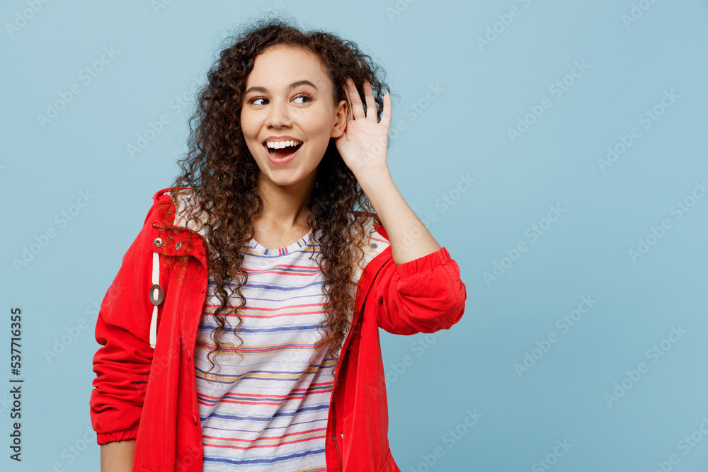 Young curious woman of African American ethnicity 20s wear red jacket try to hear you overhear listening intently isolated on plain pastel light blue cyan background. Wet fall weather season concept. Young curious woman of African American ethnicity 20s wear red jacket try to hear you overhear listening intently isolated on plain pastel light blue cyan background. Wet fall weather season concept.