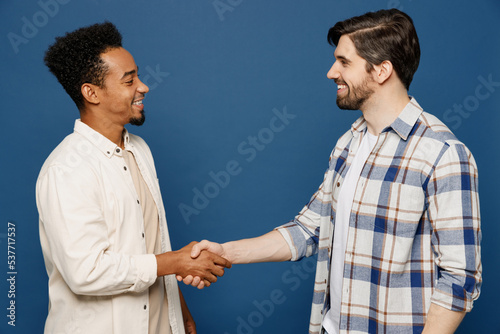 Side view young two friends cheerful men 20s wear white casual shirts looking camera together shaking hands meeting each other isolated plain dark royal navy blue background. People lifestyle concept.