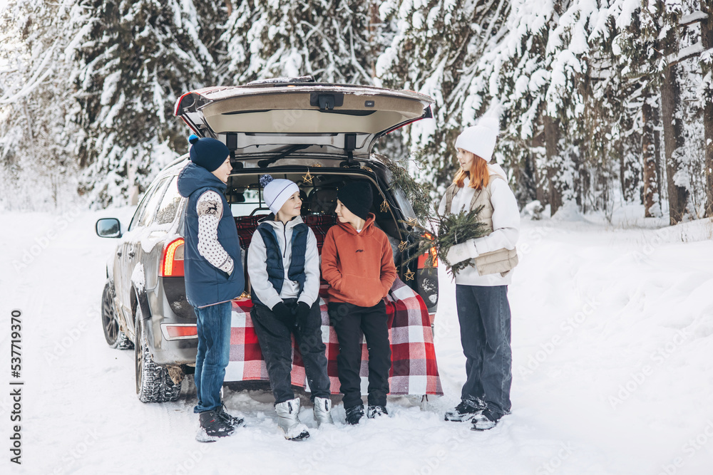Four friends teenagers boys and girl with thermos sitting in trunk of car decorated for Christmas and New year. Winter picnic in snowy forest. Road trip and local travel. Friends hanging out together
