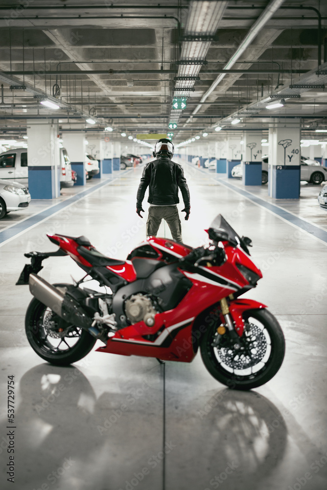 Obraz premium Back view of a motorcycle rider with a helmet and defocused race motorcycle in a car parking lot.
