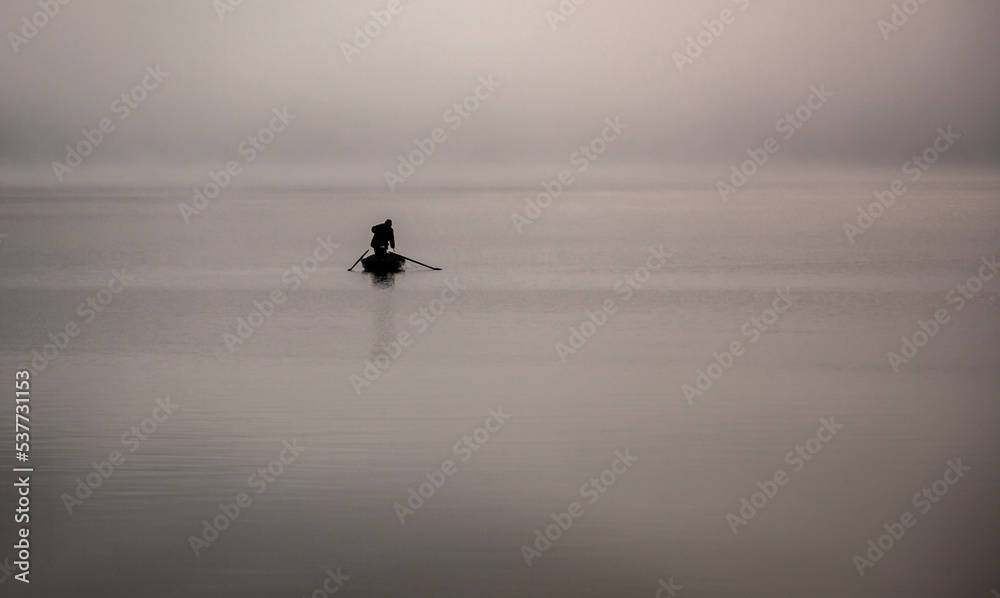Fototapeta premium Silhouette of fisherman on the water in the fog choosing fishing nets on boat with oars.