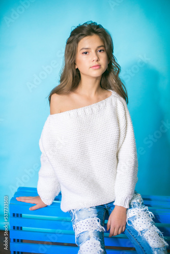 
A cute teenage girl with long dark hair in trendy jeans and a white knitted sweater on a blue background in the studio. Fashion for teenagers