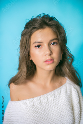 
A cute teenage girl with long dark hair in trendy jeans and a white knitted sweater on a blue background in the studio. Fashion for teenagers