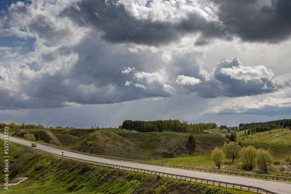 Osphalt road. Spring landscape, dramatic sky over green hills, before rain. The sun through the clouds illuminates the road and ravines.