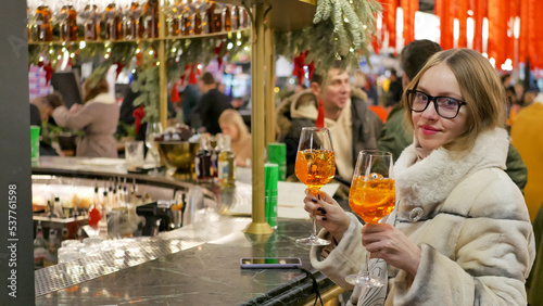 blond woman with glasses wears white winter fur coat  at the restaurant, bar, pub. Girl holds two glasses of drink in hands. Crowded place at background. Public place, celebration, having fun