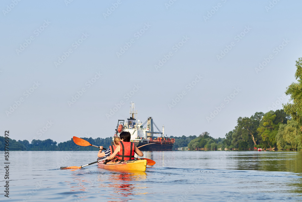 Back view of two women rows in a tandem kayak near a cargo ship anchored in Danube river at summer morning