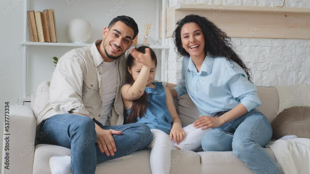 Multiracial family multiethnic Hispanic Caucasian Arabian Indian parents mother father with daughter child girl sitting on couch at home waving hands hello talking on online video call chat conference Multiracial family multiethnic Hispanic Caucasian Arabian Indian parents mother father with daughter child girl sitting on couch at home waving hands hello talking on online video call chat conference