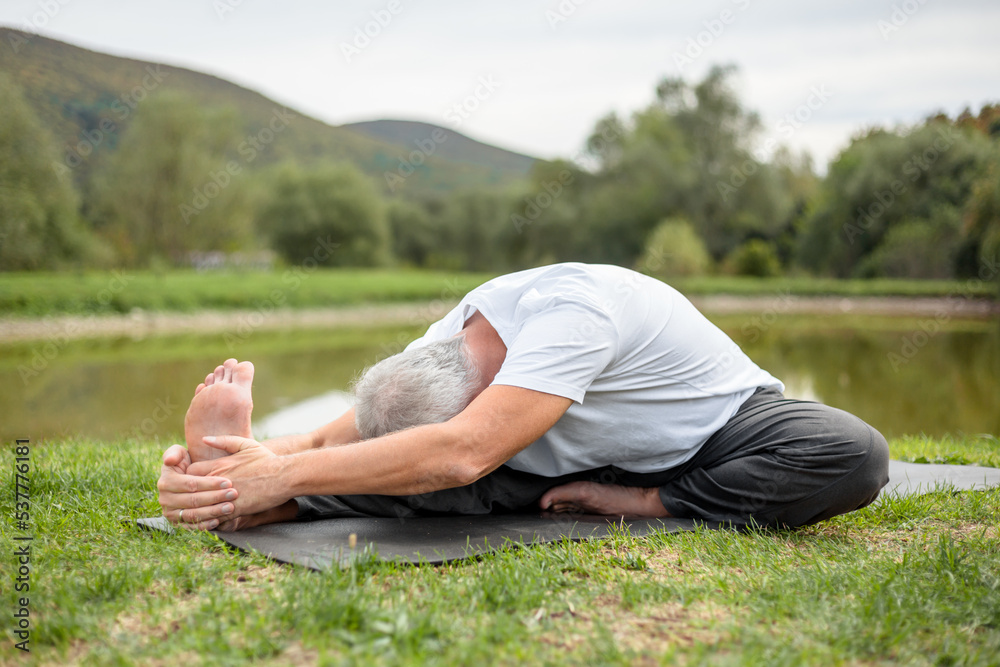 An elderly fit man is doing stretching sitting on a yoga mat in a forest clearing in the open air An elderly fit man is doing stretching sitting on a yoga mat in a forest clearing in the open air