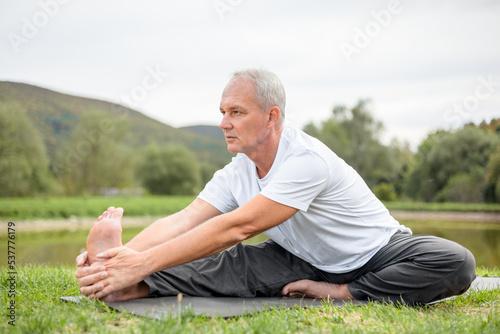 An elderly fit man is doing stretching sitting on a yoga mat in a forest clearing in the open air
