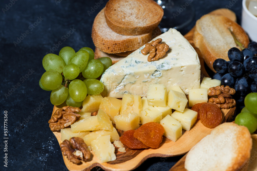 Fototapeta premium cheese platter on a wooden board, bread, fruit and cold cuts on dark background