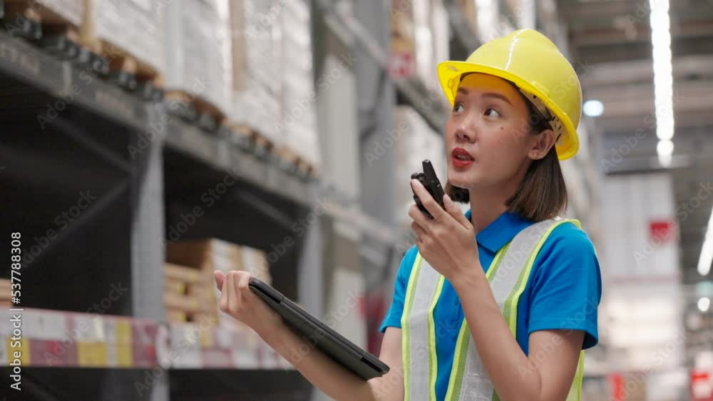 Beautiful asian engineer woman wearing safety helmet and reflective vest, She checking goods and supplies on shelves with tablet and talking to someone with walkie talkie in the warehouse. Beautiful asian engineer woman wearing safety helmet and reflective vest, She checking goods and supplies on shelves with tablet and talking to someone with walkie talkie in the warehouse.