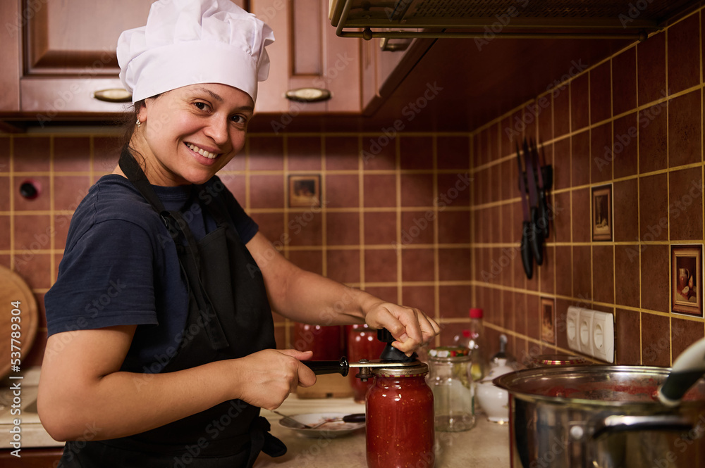 Smiling woman housewife in white chef hat and apron, using seamer, closes lids of jars with freshly canned tomato sauce, juice or passata, preserving food with organic homegrown tomatoes for winter Smiling woman housewife in white chef hat and apron, using seamer, closes lids of jars with freshly canned tomato sauce, juice or passata, preserving food with organic homegrown tomatoes for winter