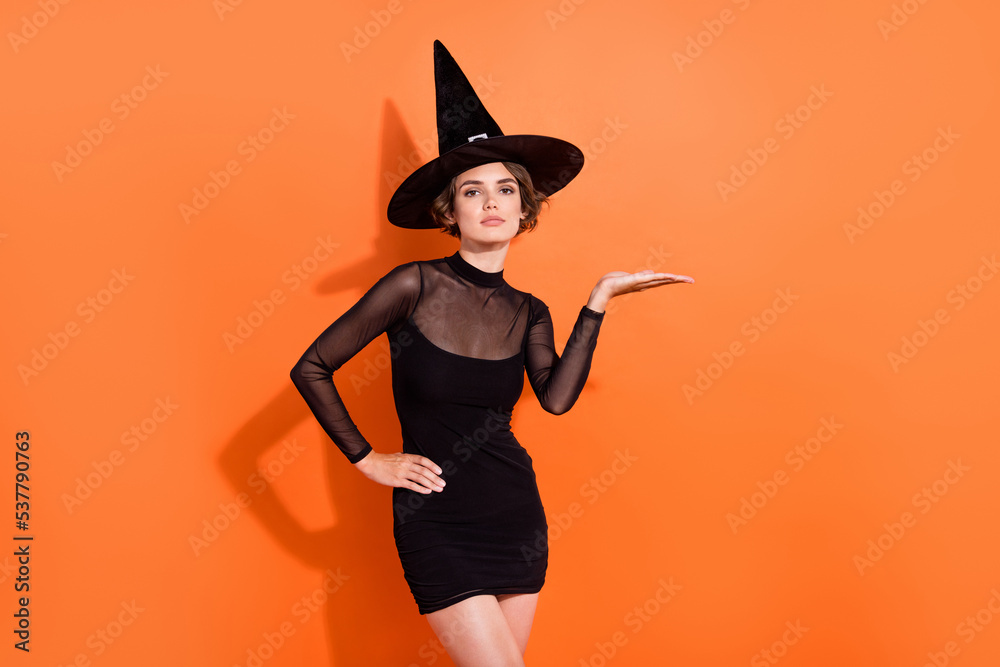 Photo of cheerful pretty lady wear black witch dress cap holding arm empty space isolated orange color background