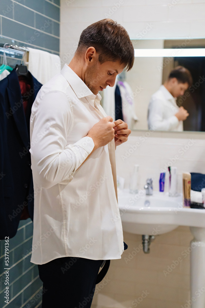 Obraz premium a man in the bathroom puts on and buttons the buttons on a white shirt. 