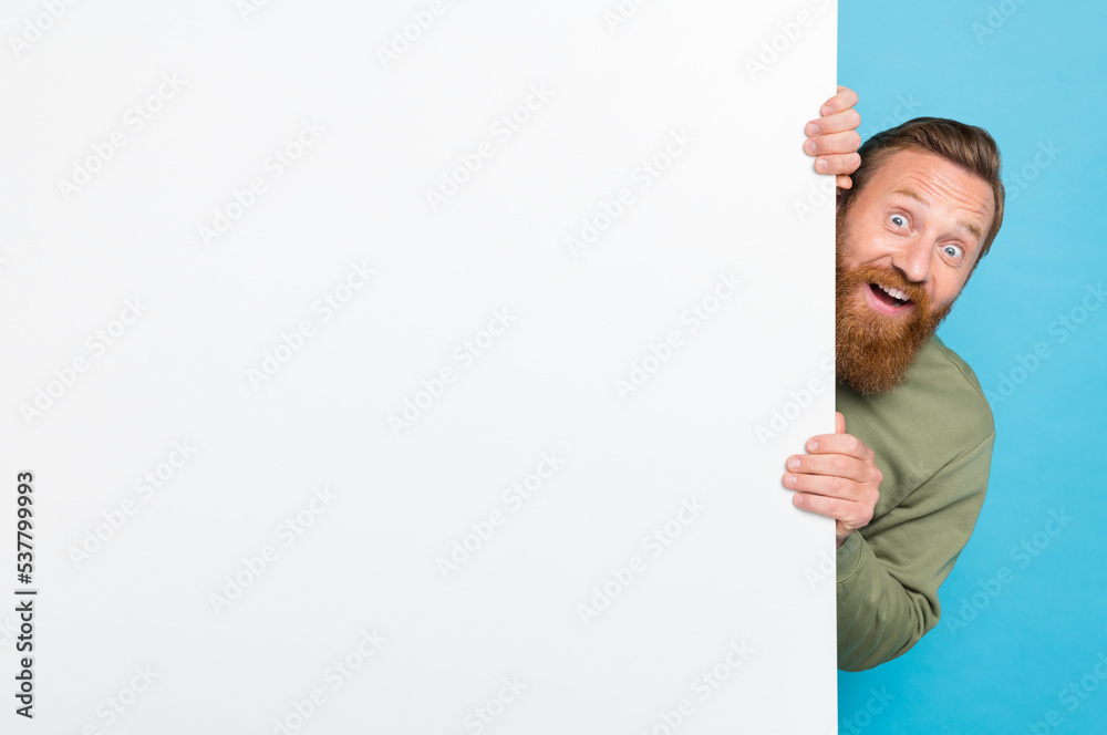 Photo portrait of nice young guy hiding behind white wall advert print space wear khaki clothes isolated on cyan color background Photo portrait of nice young guy hiding behind white wall advert print space wear khaki clothes isolated on cyan color background