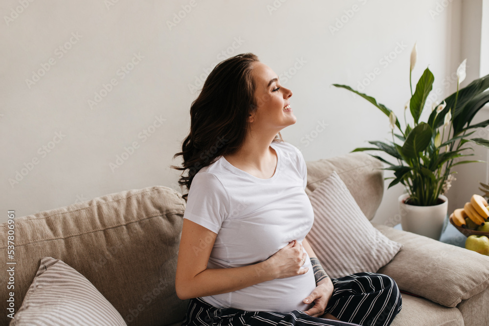 Pretty brunette curly pregnant woman in striped black pants and white tee smiles. Young mother gently touches belly and sits on sofa Pretty brunette curly pregnant woman in striped black pants and white tee smiles. Young mother gently touches belly and sits on sofa