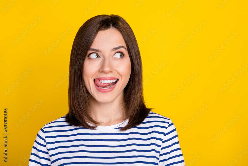 Photo portrait of gorgeous young girl licking teeth look curious empty space wear stylish striped look isolated on yellow color background Photo portrait of gorgeous young girl licking teeth look curious empty space wear stylish striped look isolated on yellow color background