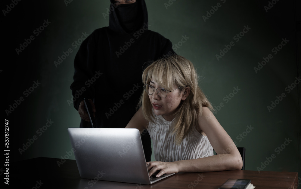 Fototapeta premium A black robed criminal with a gun at her aimed at a woman to steal her computer information. in a pitch dark room