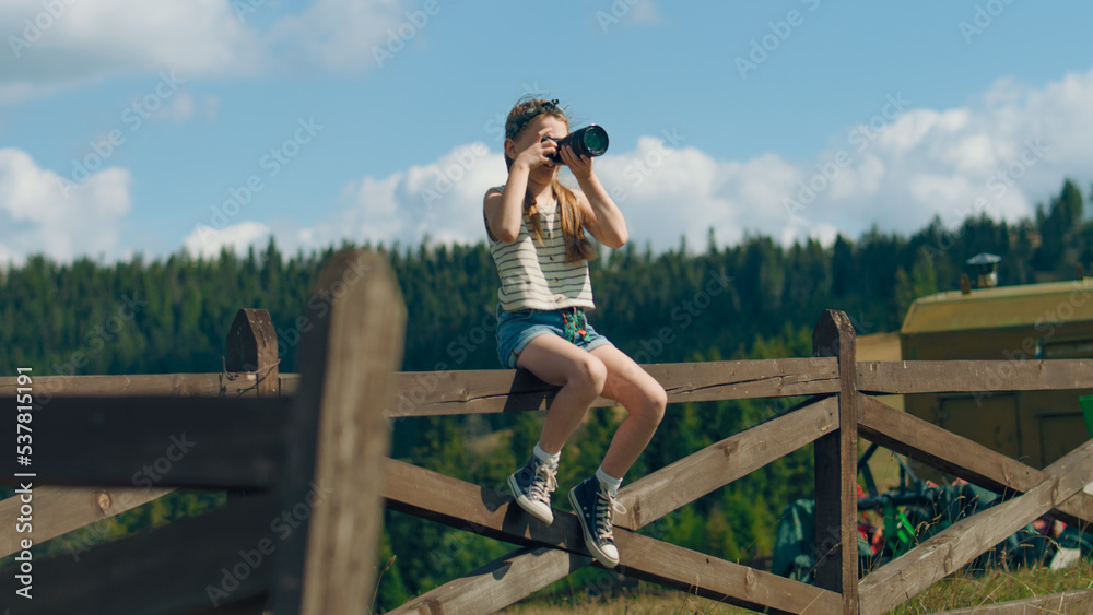 Young photographer shooting landscape using digital camera on beautifull green hillway, sitting on wooden fence, taking pictures of nature. Girl filling the portfolio with amazing photos.