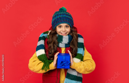 smiling teen girl wearing knitted hat hold present box on red background, xmas
