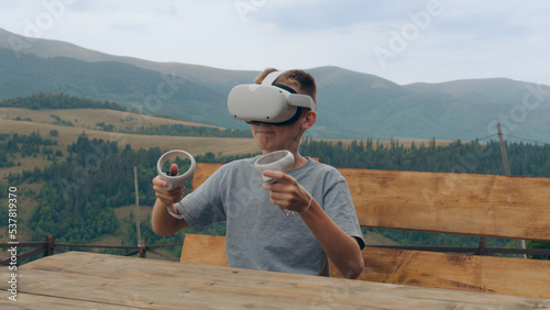 Young guy playing augmented reality games on nature, sitting on wooden bench. Gamer testing VR goggles and wireless controllers, spending leisure time at outdoor © Framestock