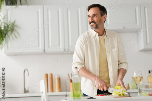 bearded man smiling and looking away while cutting lettuce near glass of white wine.