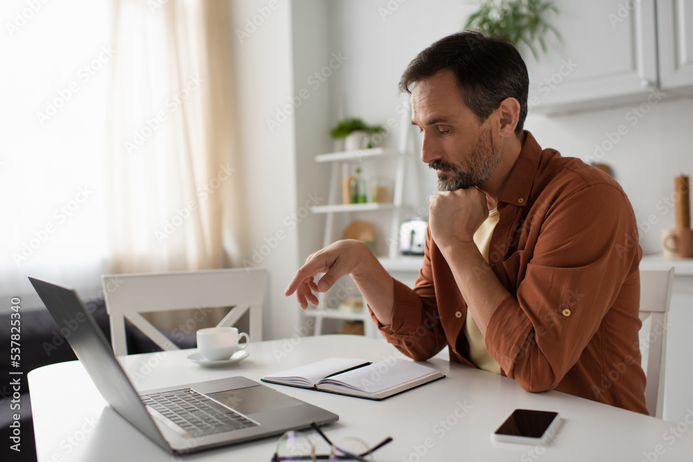 Fototapeta premium pensive man thinking near empty notebook and gadgets in kitchen.