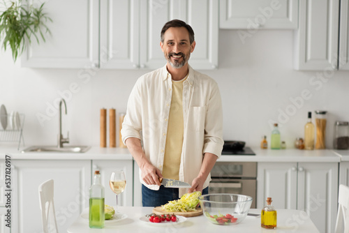 bearded man smiling at camera while preparing salad with lettuce and cherry tomatoes near glass of white wine.