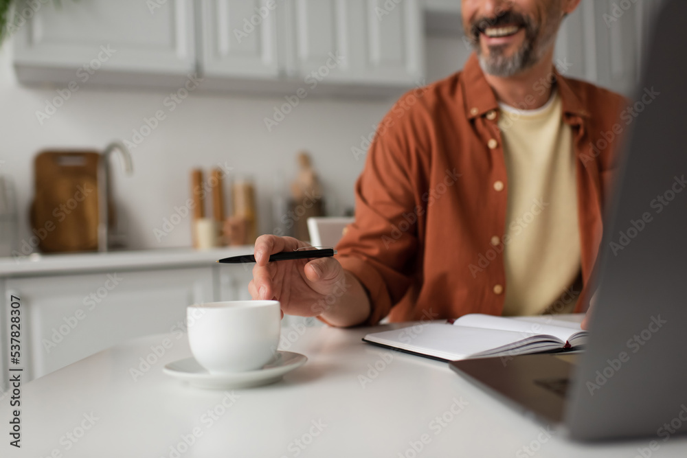 cropped view of bearded man with pen smiling near coffee cup and blurred laptop. cropped view of bearded man with pen smiling near coffee cup and blurred laptop.