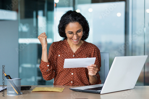 Hispanic woman received a happy letter from the bank, business woman reads and rejoices celebrating success, female worker in glasses and curly hair works inside a modern office building uses laptop. photo