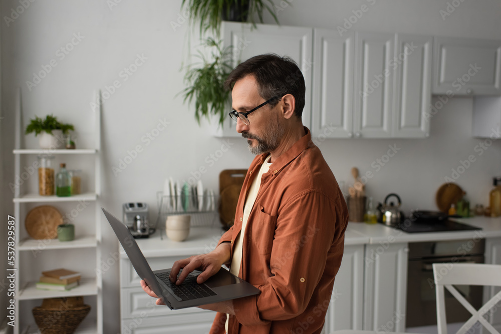 side view of bearded man in eyeglasses using laptop while standing in kitchen.