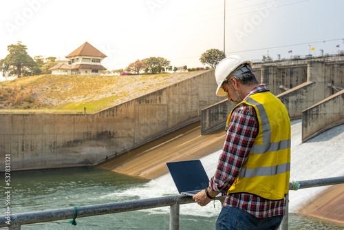 A dam engineering doing his checking routine. He is wearing a white hard hat and yellow transparent vest. He is standing by the rail by the dam.