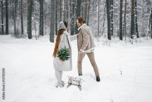 Love romantic young couple girl,guy in snowy winter forest with christmas tree, sled.Walking with sleigh in stylish clothes, fur coat,jacket, woolen shawl, bonnet.Snow lovestory.Romantic date,weekend © velirina
