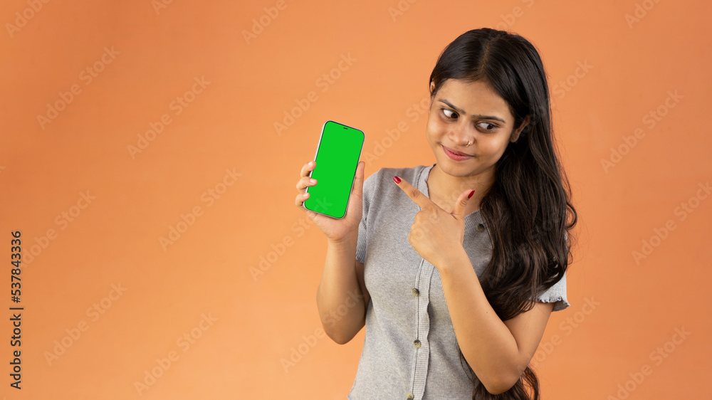Smiling young woman pointing at smartphone screen, mock up cellphone, mobile with green screen isolated over color background Smiling young woman pointing at smartphone screen, mock up cellphone, mobile with green screen isolated over color background