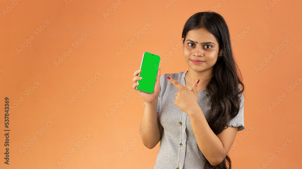 Smiling young woman pointing at smartphone screen, mock up cellphone, mobile with green screen isolated over color background