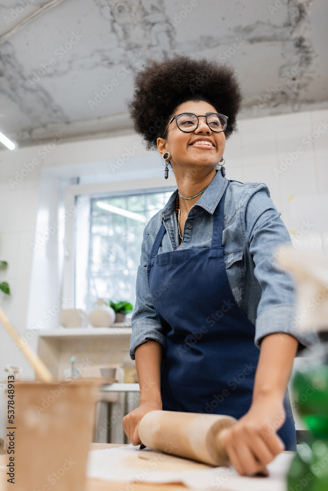 cheerful african american woman in eyeglasses and apron modeling clay with rolling pin. cheerful african american woman in eyeglasses and apron modeling clay with rolling pin.
