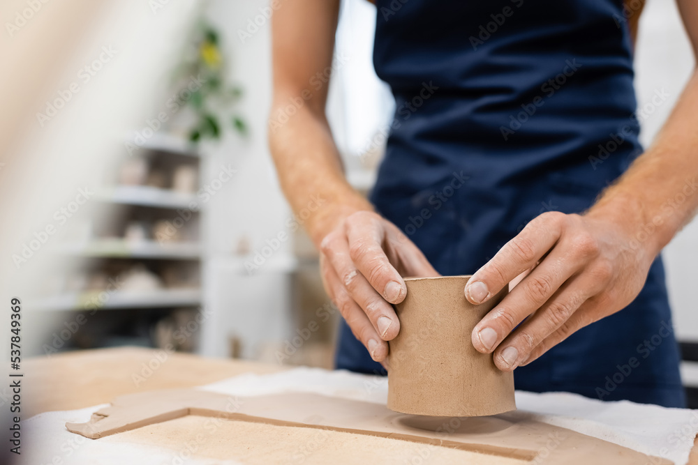cropped view of man in blue apron molding clay cup in pottery workshop.