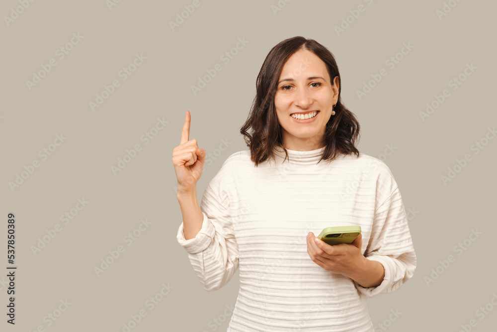Young smiling woman points above up to copy space while holding her phone. Young smiling woman points above up to copy space while holding her phone.