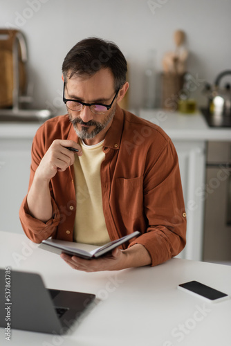 pensive man in eyeglasses looking at notebook near blurred laptop and mobile phone with blank screen.