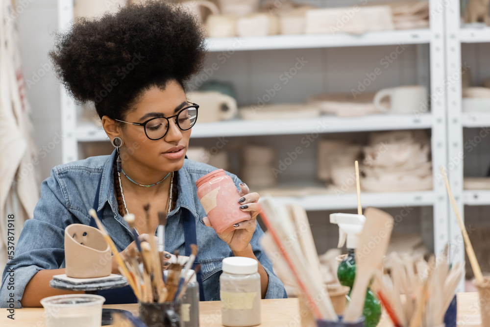 Fototapeta premium African american craftswoman in eyeglasses holding jar with paint near blurred paintbrushes and clay sculpture in pottery studio.