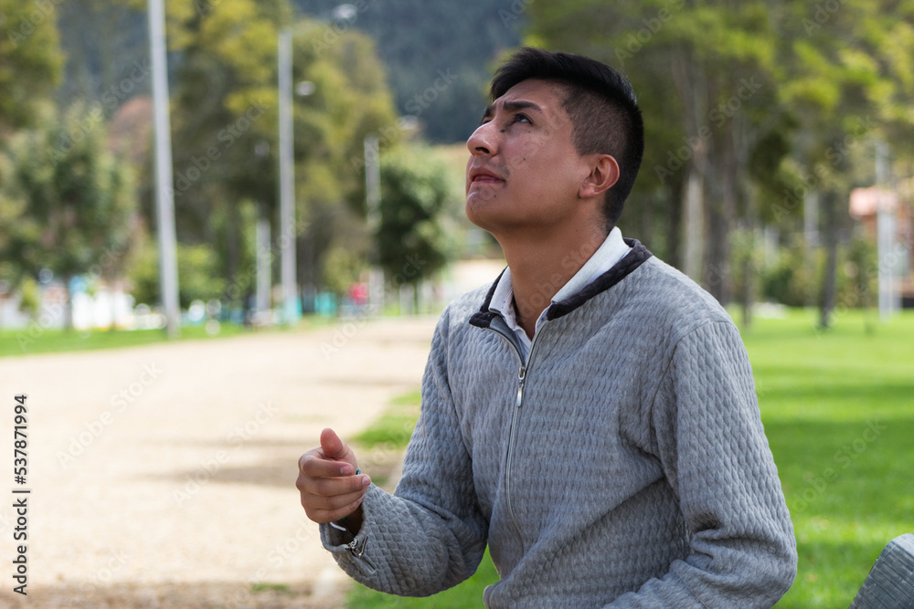 A repentant young Latin man asking for forgiveness sitting in the middle of a park on a sunny day A repentant young Latin man asking for forgiveness sitting in the middle of a park on a sunny day